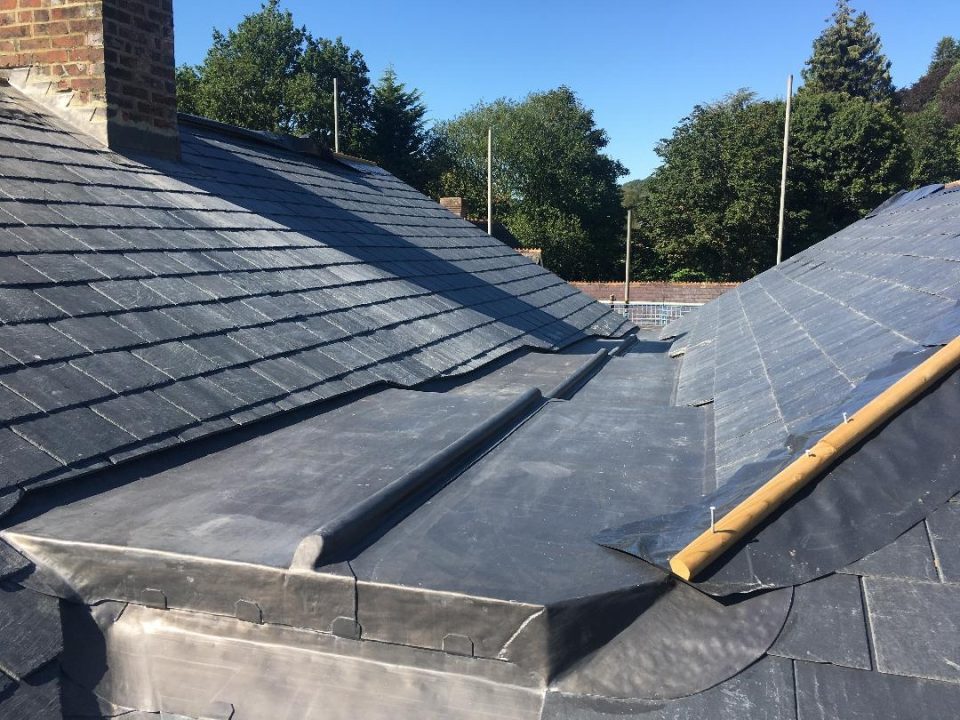 A rooftop covered with slate tiles features a lead valley for drainage. The lead valley is positioned between two sloped sections of the roof. The surrounding area includes trees, with some scaffolding poles visible in the background. The weather is clear and sunny.